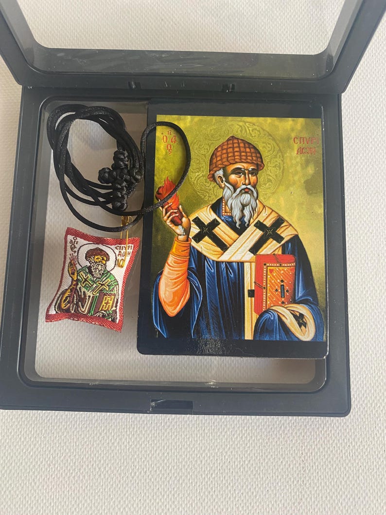 May include: A religious icon and pendant set in a black display case. The icon depicts a saint holding a flame and a book, with text in a foreign language. A small pendant with a similar image hangs from a black cord.