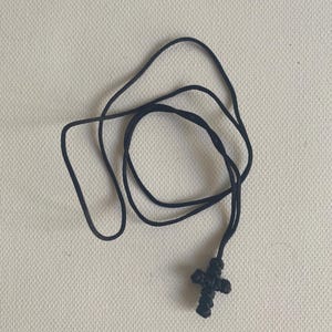 May include: A black cross pendant necklace. The cross is made of woven material and is attached to a black cord. The necklace is displayed on a neutral background.
