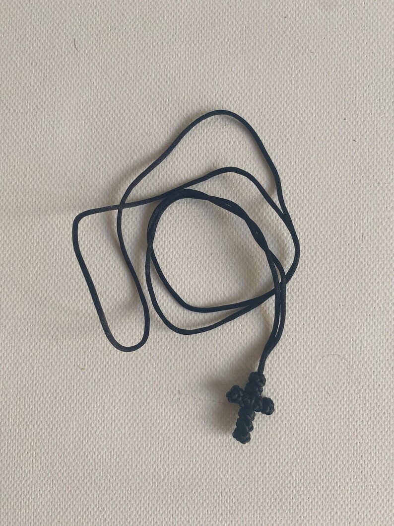 May include: A black cross pendant necklace. The cross is made of woven material and is attached to a black cord. The necklace is displayed on a neutral background.