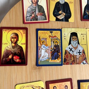 May include: A collection of religious icon cards featuring various saints and religious figures. The cards have colorful illustrations with gold accents and text in a foreign language. The cards are arranged on a light brown wooden surface.