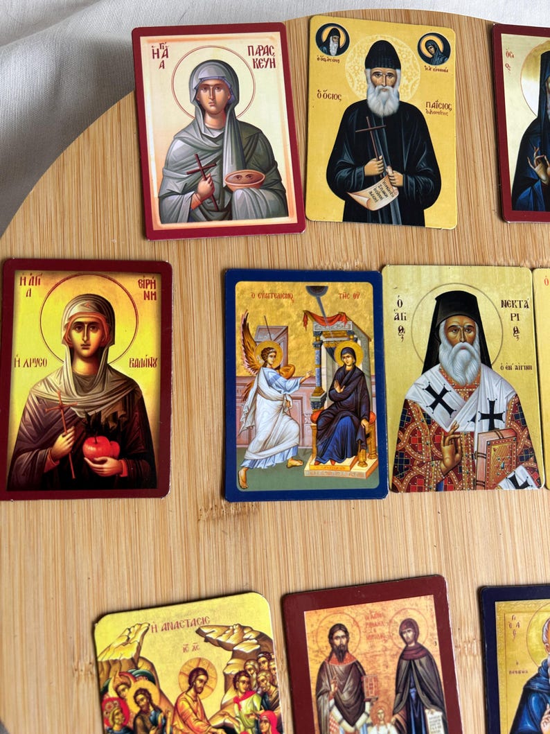 May include: A collection of religious icon cards featuring various saints and religious figures. The cards have colorful illustrations with gold accents and text in a foreign language. The cards are arranged on a light brown wooden surface.
