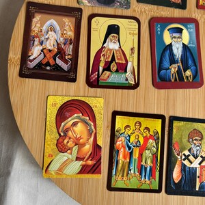 May include: A collection of religious icon cards featuring various saints and biblical scenes. The cards display vibrant colors and intricate details, with gold accents and ornate borders. The images are set against a light wooden background.