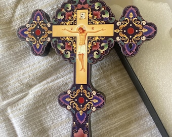 May include: A wooden cross with a purple and gold floral design. The centre of the cross features a depiction of Jesus Christ on the cross. The cross is decorated with red and gold accents.