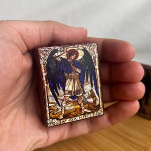 May include: A rectangular wooden plaque featuring a detailed illustration of St. Michael, with a halo, wings, and a staff, standing over a serpent. The text "ST MICHAEL" is at the bottom. The artwork has a stained-glass style.