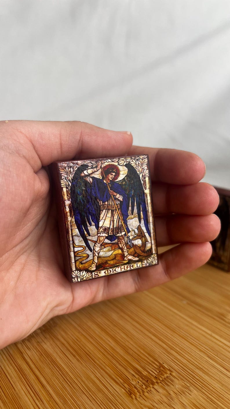May include: A rectangular wooden plaque featuring a detailed illustration of St. Michael, with a halo, wings, and a staff, standing over a serpent. The text "ST MICHAEL" is at the bottom. The artwork has a stained-glass style.