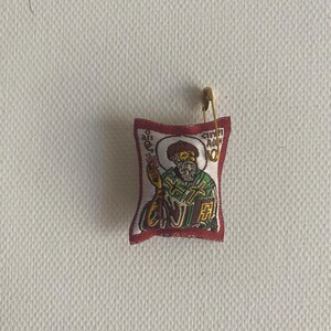 May include: A small, rectangular religious icon with a gold safety pin attached. The icon features an image of a saint in green and gold robes, with text in a foreign language. The border is red.