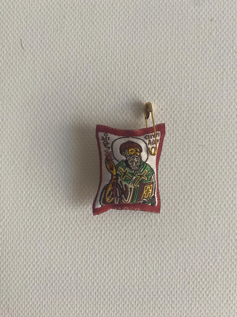 May include: A small, rectangular religious icon with a gold safety pin attached. The icon features an image of a saint in green and gold robes, with text in a foreign language. The border is red.