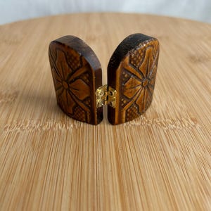 May include: A small, two-part wooden object with a dark brown finish, featuring carved floral patterns and a cross-hatched texture. The two pieces are connected by a small, gold-coloured metal hinge in the centre, allowing them to open and close.