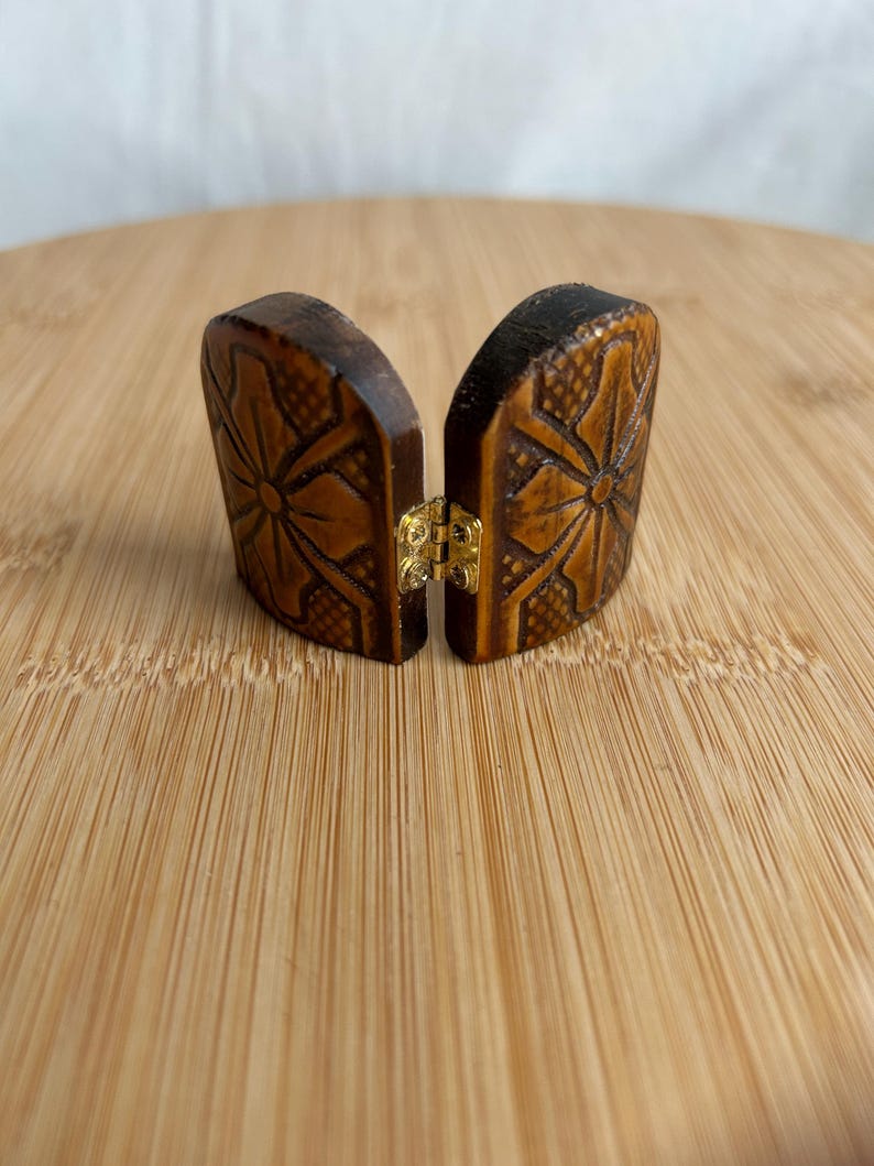 May include: A small, two-part wooden object with a dark brown finish, featuring carved floral patterns and a cross-hatched texture. The two pieces are connected by a small, gold-coloured metal hinge in the centre, allowing them to open and close.