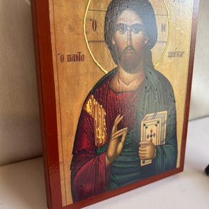 Może przedstawiać: Tradycyjna ikona religijna przedstawiająca Jezusa Chrystusa trzymającego książkę. Ikona jest namalowana na drewnianym panelu z czerwoną ramą. Tło jest jasnobrązowe z złotymi akcentami. Tekst na ikonie brzmi "&Omicron; &Pi;&Alpha;&Nu;&Tau;&Omicron;&Kappa;&Rho;&Alpha;&Tau;&Omega;&Rho;", co tłumaczy się jako "Wszechmogący".