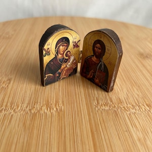 May include: A small, hinged wooden diptych icon featuring religious imagery. The left panel depicts a figure holding a child against a gold background, while the right panel shows a bearded figure holding a book, also set against a gold background.
