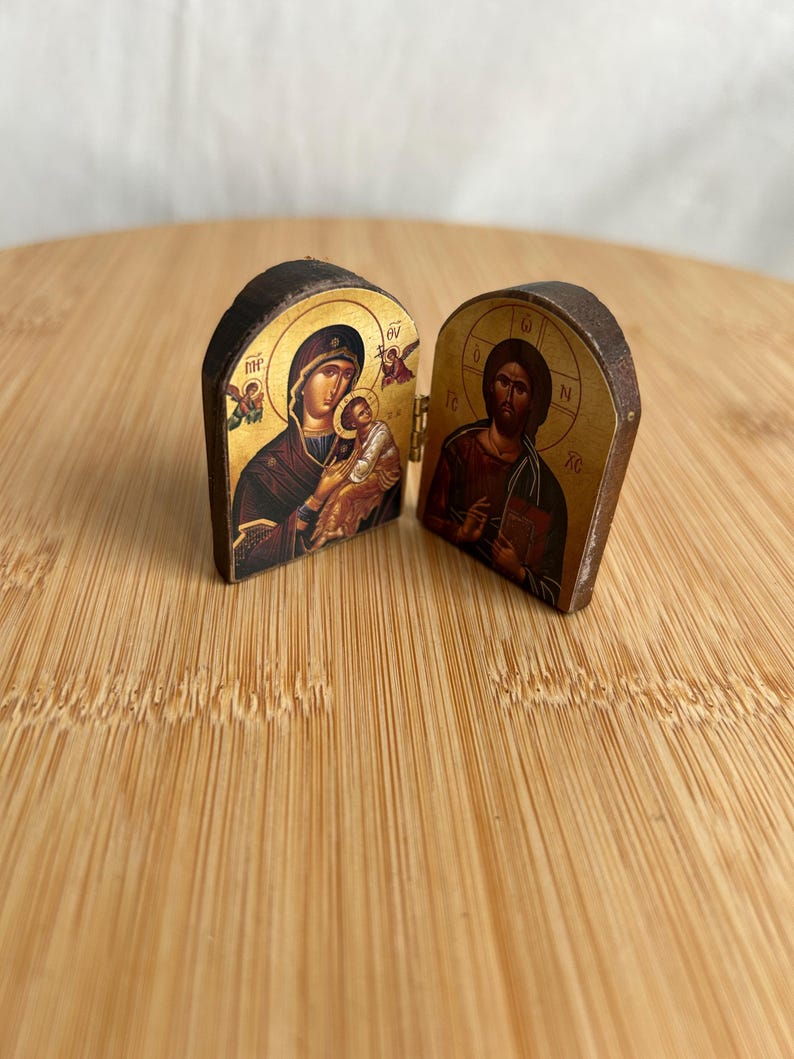 May include: A small, hinged wooden diptych icon featuring religious imagery. The left panel depicts a figure holding a child against a gold background, while the right panel shows a bearded figure holding a book, also set against a gold background.