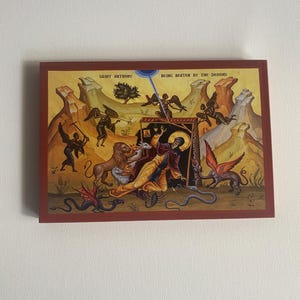 以下が含まれることがあります： 悪魔に打ちのめされる聖アントニウスを描いた、額装された宗教的なイコン。この作品は、さまざまな生き物や人物に囲まれた中心人物を特徴とし、上部には「Saint Anthony Being Beaten By The Demons」と書かれています。