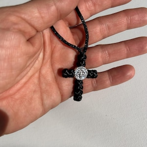 May include: A black cord necklace with a silver cross pendant. The cross is made of braided cord and has a silver centre piece with a floral design.