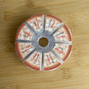 May include: A circular, clear plastic container divided into seven wedge-shaped compartments, each filled with small, light-colored solid pieces. Each section features a label with text in English and Greek: Nightflower, Jasmine, Manolia, Rose, Byzantine, Honeysuckle, and Desert Flower, along with Nard.