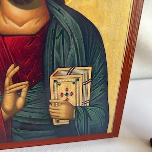 Może przedstawiać: Ikona religijna przedstawiająca Jezusa Chrystusa trzymającego książkę z napisem "KPATOQ" nad nim. Ikona jest namalowana w tradycyjnym stylu z żywymi kolorami i misternymi detalami.