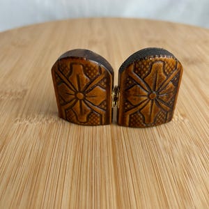 May include: A pair of brown, arched, semi-circular wooden pieces joined by a small gold-toned metal hinge. Each piece features a carved, symmetrical floral pattern with a central circular element and a textured, cross-hatched background design.