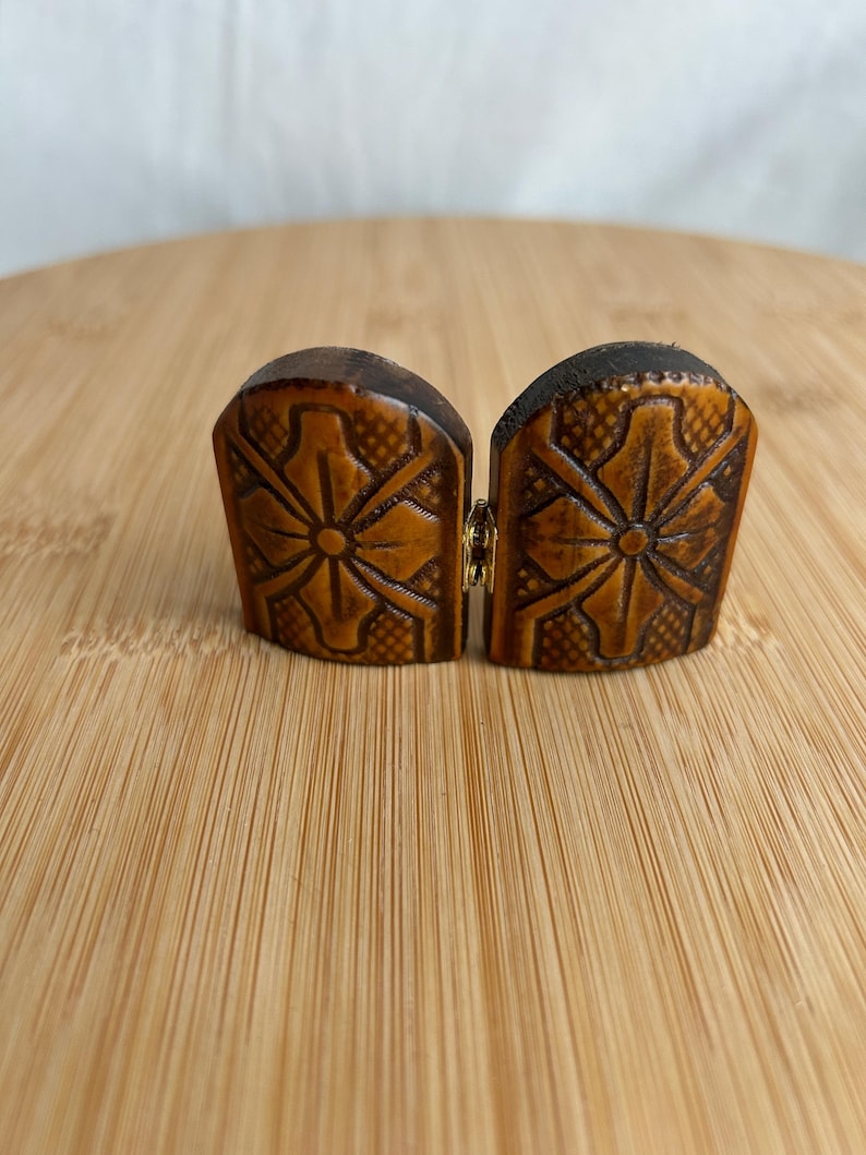 May include: A pair of brown, arched, semi-circular wooden pieces joined by a small gold-toned metal hinge. Each piece features a carved, symmetrical floral pattern with a central circular element and a textured, cross-hatched background design.