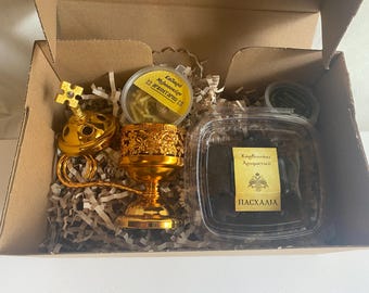 May include: A gold-colored censer, incense, and other religious items are arranged inside a cardboard box. The box contains a clear container with a label, a small round container, and decorative paper.