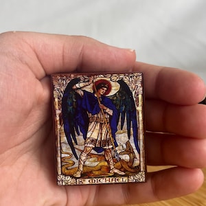 Könnte beinhalten: Rechteckiges Bild im Glasmalerei-Stil von St. Michael, der einen Drachen tötet. Das Kunstwerk zeigt eine Figur in blauen und weißen Gewändern mit rotem Heiligenschein, die einen Speer hält. Der Text "ST MICHAEL" steht unten.