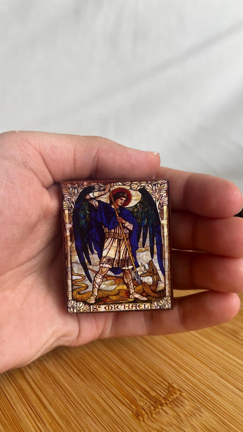 May include: A rectangular, stained-glass style image of St. Michael slaying a dragon. The artwork features a figure in blue and white robes with a red halo, holding a spear. The text "ST MICHAEL" is at the bottom.