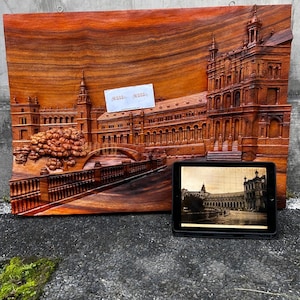 May include: A rectangular wooden wall plaque featuring a detailed relief carving of the Plaza de España in Seville, Spain. The carving depicts the architectural arches, towers, and a bridge. A small white card with text is attached to the surface.