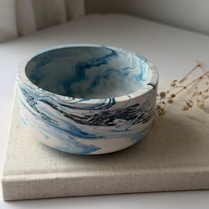 May include: A round, shallow bowl featuring a marbled pattern of white, deep blue, and black swirls. The exterior and interior surfaces display a smooth, continuous finish with fluid, organic colour transitions throughout the entire vessel.