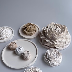 May include: A selection of floral-shaped ornaments in varying sizes and shades. The objects are displayed on circular, off-white trays and a light grey surface. The flowers range in colour from white to beige and brown.