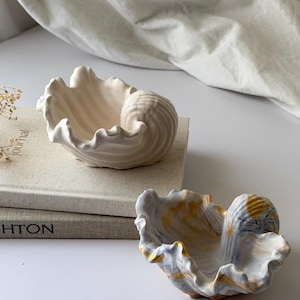 May include: Two ceramic shell-shaped bowls. One is solid off-white, the other has a marbled design in white, gold, and blue. The bowls have a textured, wavy rim and a spiral-shaped centre, perfect for decorative use.