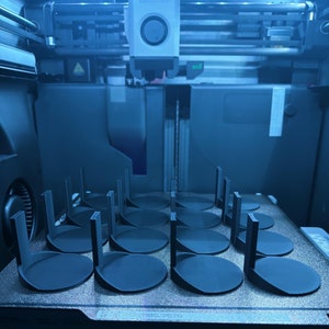 May include: A close-up of a 3D printer bed with 12 printed circular objects. The objects are arranged in a grid pattern and are made of a dark gray material. The objects are all the same size and shape, and they have a smooth, rounded surface.
