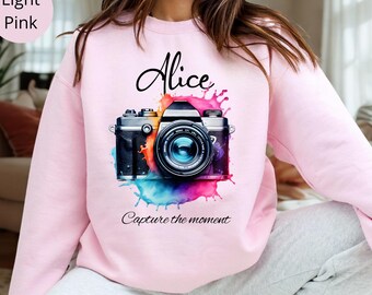 May include: Light pink sweatshirt featuring a colorful watercolor design of a vintage camera. The name "Alice" is written above the camera, with the phrase "Capture the moment" below. The sweatshirt is a solid light pink color.