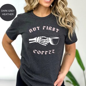 May include: Dark grey heather t-shirt with the text "But First Coffee" in a light pink font. The graphic features a skeleton hand holding a coffee cup. The shirt is short-sleeved.