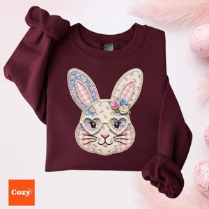 Può includere: Felpa bordeaux con un'applicazione a forma di coniglio. Il coniglio ha occhiali a forma di cuore, decorazioni floreali e un motivo a quadretti. La parola "Cozy" è nell'angolo in basso a sinistra. Perfetta per Pasqua o la primavera.