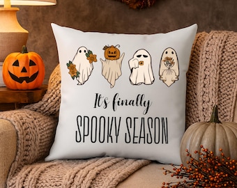 May include: White square pillow with the text "It's finally SPOOKY SEASON" in black. The pillow features four cartoon ghosts with floral and pumpkin accents. A carved pumpkin and a decorative gourd are also visible.