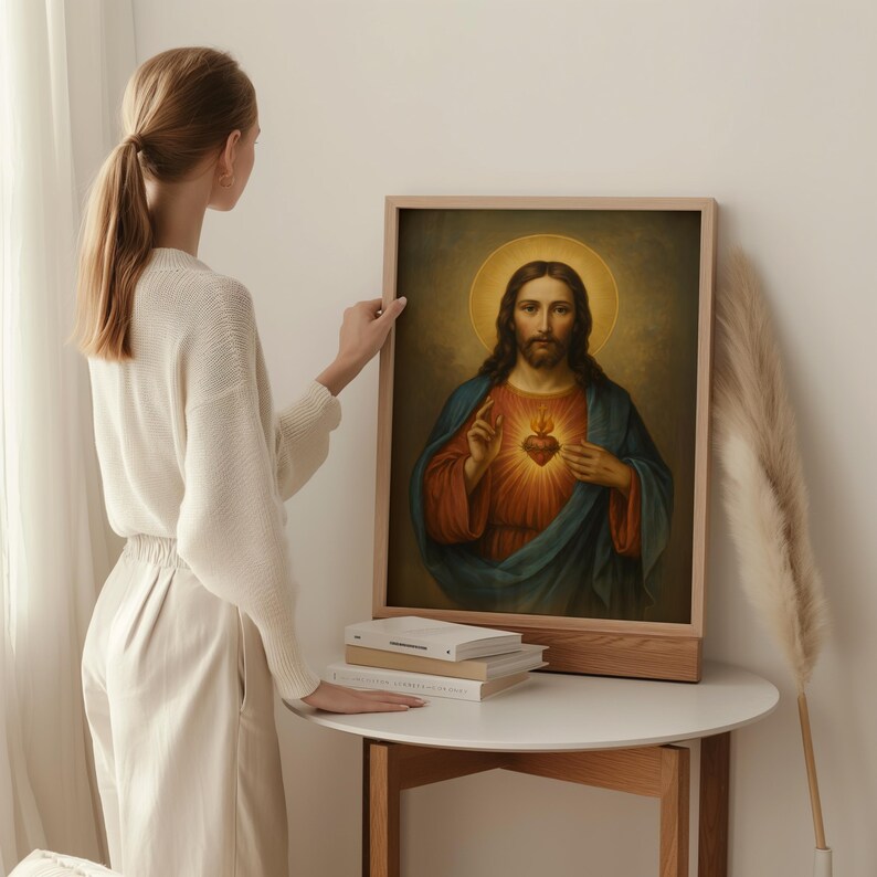 May include: A framed religious portrait of Jesus Christ, featuring a golden halo, a red heart, and blue and red robes. The artwork is displayed on a wooden stand, with a stack of books on a white table. The image is set against a neutral background.