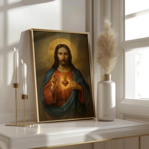 May include: A framed religious portrait featuring a figure with long brown hair, a halo, and a red heart. The figure wears a blue robe and a red tunic. The gold frame rests on a white surface, next to a vase with pampas grass and gold candle holders.