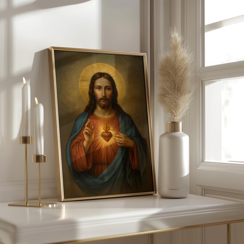 May include: A framed religious portrait featuring a figure with long brown hair, a halo, and a red heart. The figure wears a blue robe and a red tunic. The gold frame rests on a white surface, next to a vase with pampas grass and gold candle holders.