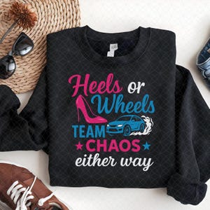 May include: Black crew neck sweatshirt with the text "Heels or Wheels TEAM CHAOS either way." The design includes a pink high heel and a blue car. The text and graphics are in pink, blue, and white.