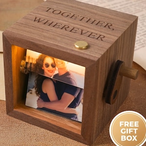 以下が含まれることがあります： 「TOGETHER, WHEREVER」の文字が刻まれた木製のフォトキューブ。小さな真鍮製のボタンと回転式のハンドルが付いています。キューブの中にはカップルの写真が見えます。キューブは温かみのある茶色です。