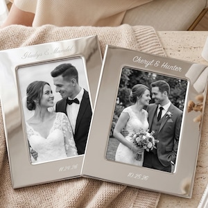 May include: Two rectangular silver-toned metal picture frames with engraved text. The left frame is engraved with Gladys & Mandel and 11.24.26. The right frame is engraved with Cheryl & Hunter and 10.25.26. Each frame holds a black and white wedding photo.