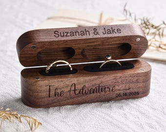 May include: A wooden ring box with two compartments for rings. The box is engraved with the text "Suzanah & Jake" and "The Adventure 06.16.2025".