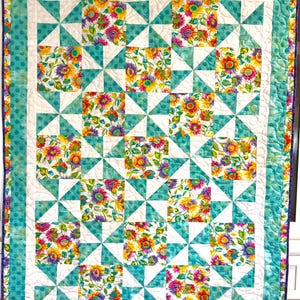 May include: A rectangular quilt featuring a repeating pattern of floral squares with sunflowers and purple blossoms, alternating with white and teal pinwheel blocks. The quilt is framed by a white border and an outer teal polka dot border.