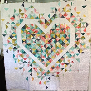 May include: A white quilt with a large heart-shaped design made of colorful patchwork triangles. The heart is surrounded by a white border and the quilt is made of a variety of fabrics with different patterns.