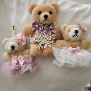 Puede incluir: Tres osos de peluche de color marrón claro. Los osos están adornados con lazos, encajes y faldas de tul en tonos rosa y blanco. Un oso tiene una pieza central decorativa con joyas. Adornos decorativos.