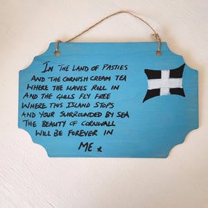 May include: A blue wooden sign with a decorative shape and a jute string for hanging. The sign features black text with a poem about Cornwall, England, and a painted black and white cross. The sign is a decorative item.