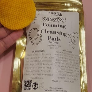 Könnte beinhalten: Ein goldener Folienbeutel mit "TURMERIC Foaming Cleansing Pads" und einem gelben, strukturierten Pad. Das Etikett listet Zutaten und Anweisungen auf. Das Pad wird in der Nähe des Beutels gehalten und zeigt seine waffelartige Textur. Das Produkt wird in den USA handgefertigt.