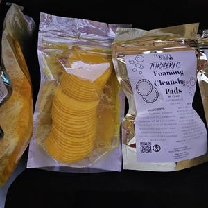 Puede incluir: Se muestran varios paquetes de productos para el cuidado de la piel. Uno contiene almohadillas amarillas circulares y otros contienen una sustancia gelatinosa amarilla. Los paquetes tienen etiquetas que dicen "Turmeric Foaming Cleansing Pads".
