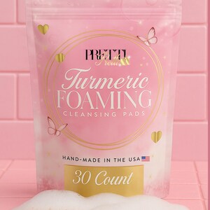 Può includere: Confezione rosa di "PRETTI" Turmeric Foaming Cleansing Pads, con dettagli dorati, farfalle e cuori. Il testo "HAND-MADE IN THE USA" e "30 Count" sono visibili. I cuscinetti sono gialli con schiuma bianca.