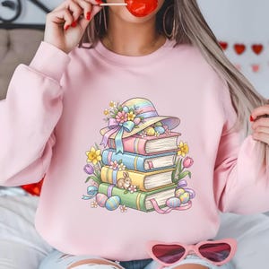 May include: Pale pink sweatshirt with an Easter-themed design. A stack of books is topped with a pastel hat decorated with flowers and Easter eggs. A bunny and more eggs are scattered around the books. The design uses a pastel colour palette.
