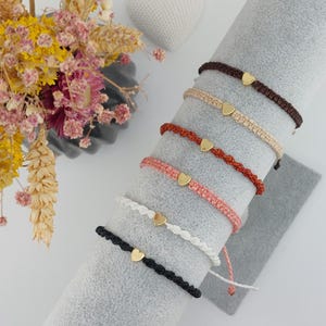 Könnte beinhalten: Eine Auswahl an handgefertigten Makramee-Armbändern in verschiedenen Farben, darunter Schwarz, Weiß, Rosa, Rot und Braun. Jedes Armband hat einen kleinen goldenen Herzanhänger. Die Armbänder werden auf einer grauen zylindrischen Oberfläche präsentiert.
